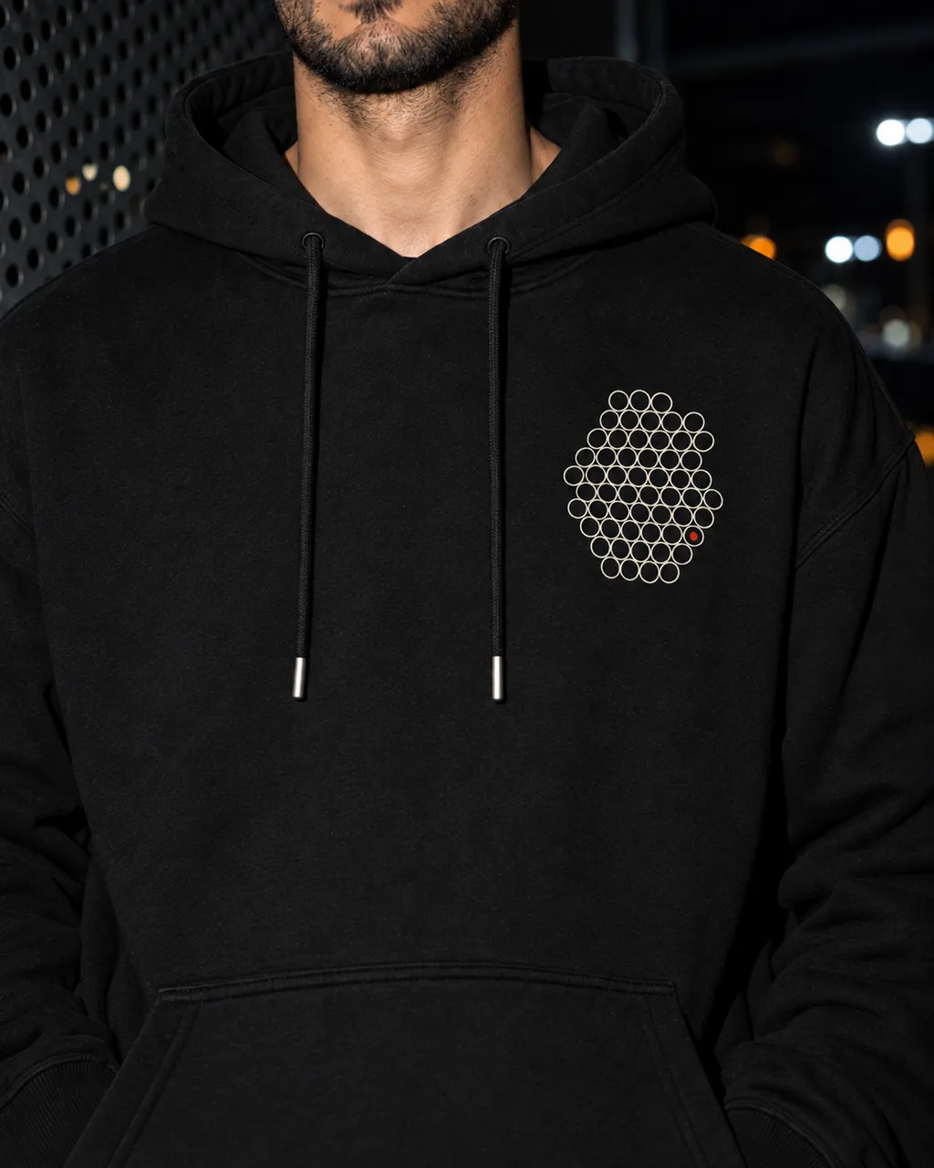 Gözenek Premium Siyah Hoodie: göğüsteki baskı yakından, gece delikli metal bir cephenin önünde