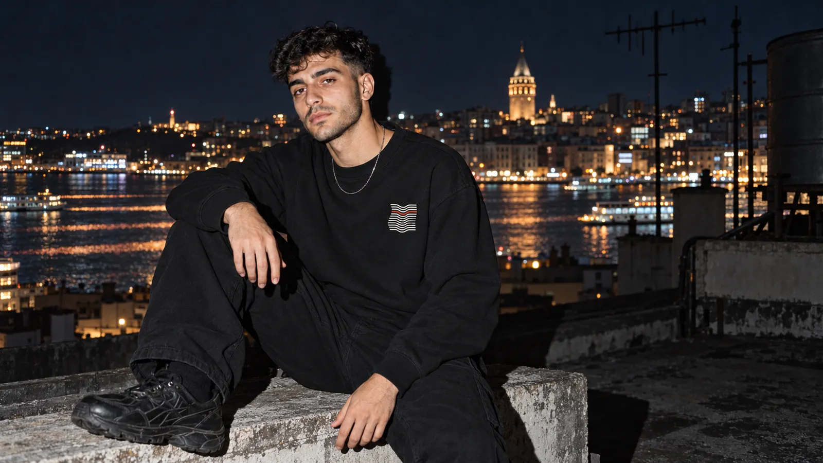Kıvrım desenli siyah sweatshirt giyen bir model, gece Boğaz'a bakan bir çatıda