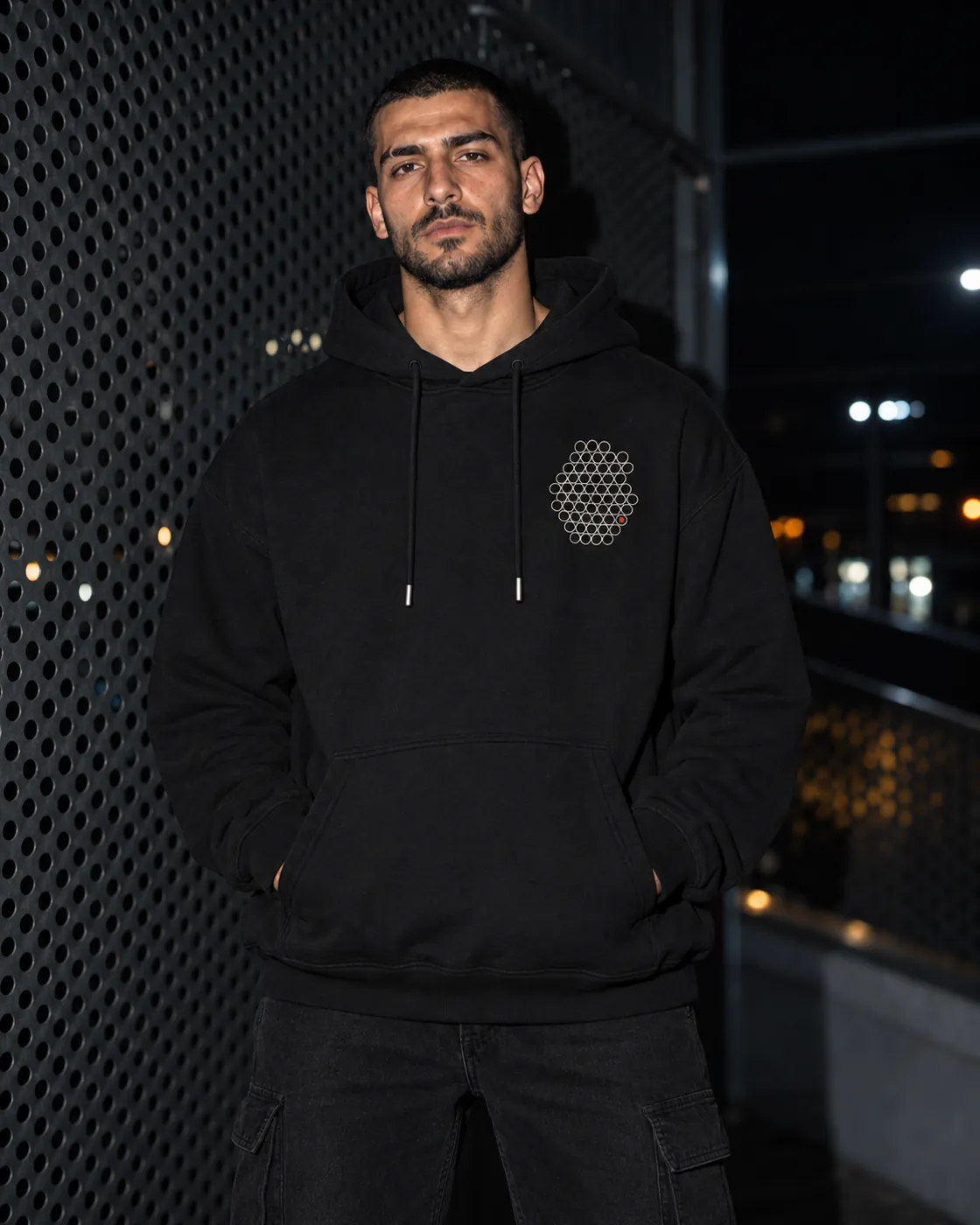 Gözenek Premium Siyah Hoodie: model üzerinde, gece delikli metal bir cephenin önünde